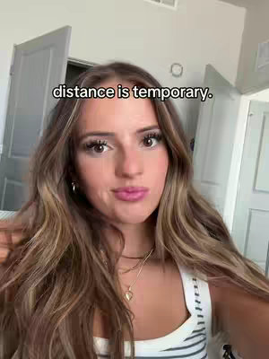 A young woman with long, wavy brown hair, wearing a white and black striped top, looks directly at the camera. Text overlayed on the image reads "distance is temporary."