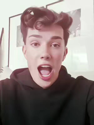 A close-up shot of a person with light skin, dark hair styled with a voluminous wave. The person, with open mouth and seemingly surprised expression, is wearing a black hoodie. The background appears to be a room with artwork on the wall.