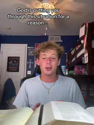 A young man with light skin and short, light-brown hair is centered in the image. He is in a room with a lit ceiling fan and a white door in the background. He is wearing a light blue t-shirt and a gold cross necklace as he holds an open book in his hands, which is likely a Bible. Behind him, various items suggest a bedroom scene, including artwork and a desk.