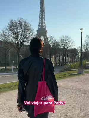 A person with their back to the camera, holding a pink tote bag, walks on a path. In the background, the Eiffel Tower is visible. The scene is set on a sunny day.