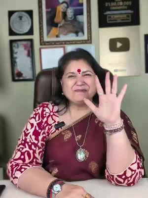 A woman with dark hair and a red bindi on her forehead smiles, holding up her right hand with five fingers extended. She is wearing a maroon and patterned red top and a necklace with a pendant. Behind her are framed pictures and a gold YouTube play button plaque. She is in what appears to be a home office setting.
