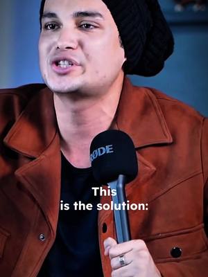 A close-up shot of a man holding a microphone. He is wearing a black beanie and a brown jacket over a black shirt. The microphone has a black cover, and the text 'This is the solution:' is overlaid on the image.