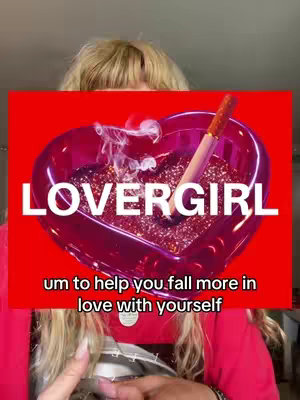 The image features a woman partially visible in the foreground, with blonde hair framing her face. A red background dominates the scene, overlaid with a heart-shaped dish filled with glitter and a cigarette. The text "LOVERGIRL" is prominently displayed above the heart, and smaller text appears below.