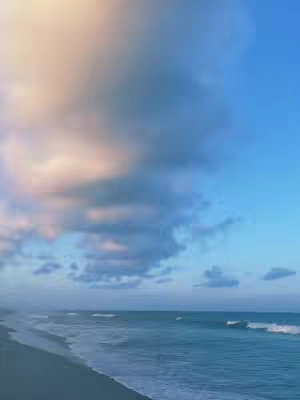 The image shows a coastal scene. The sky transitions from a peachy orange at the top left to a deep blue on the right. Thick, dark clouds dominate the center, slightly obscuring the horizon. Below the cloud line is the ocean, with waves visible. A dark beach extends along the bottom edge.