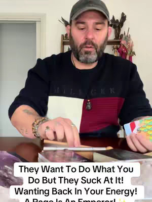 The cover image is a medium shot of a man in a room. The man is wearing a black and burgundy striped sweater with a navy baseball cap; he has a beard and mustache. He appears to be holding and handling tarot cards on a table in front of him. The image has a warm and personal feel, with a slightly blurred background suggesting depth and focus on the subject's actions. The image is a still from a video.
