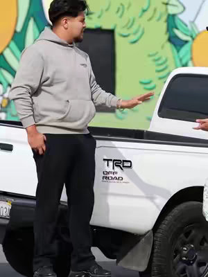 A man in a gray hoodie and black pants stands next to a white pickup truck. The truck has a black bed and the text "TRD OFF ROAD" printed on the side. The man is gesturing with his right hand, and the background includes a colorful mural.