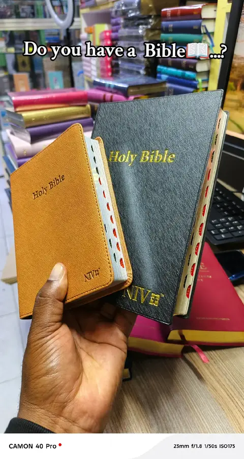 A close-up shot shows a hand holding two Bibles against a backdrop of bookshelves. The Bibles are open, displaying their pages alongside each other. The hand is the central focus, and the setting looks like a bookstore or a similar retail environment.