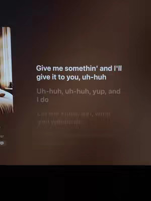 The image is a dark, moody shot of a screen displaying song lyrics. The background is a blurred, warm gradient, possibly depicting a bedroom with the corner of a window and curtains visible on the left. The focus is on the centered, white text overlaying the image. The lyrics are phrases from a song.