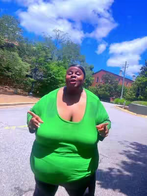 A plus-size Black woman stands on a paved road, arms outstretched and pointing outwards in each direction. She wears a vibrant green top and dark pants. The background consists of greenery and a bright blue sky with fluffy white clouds.