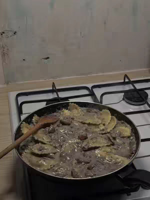 A pan of ravioli with a creamy sauce is on a stovetop. A wooden spoon rests alongside the pasta. The background consists of a white wall and the top of the stove.