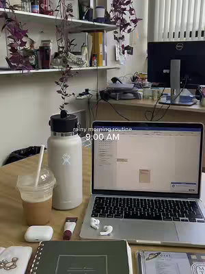 An inviting workspace, with a beige Hydro Flask, a laptop displaying a spreadsheet, a clear drink in a plastic cup, next to a journal, a pair of white earbuds, and other accessories arranged on a wooden desk, against a backdrop of shelves and purple hanging plants. The image evokes a sense of calm and routine, possibly signaling a productive morning.