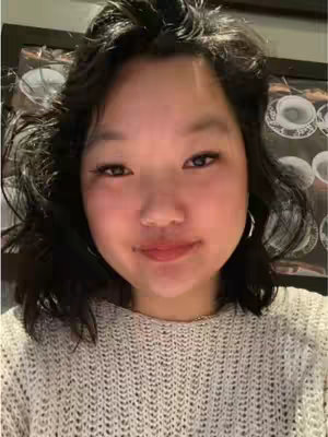A selfie-style shot featuring a young woman with dark hair, wearing a light-colored knit sweater. She has dark eyes and a slightly upward gaze, with a subtle smile. The background appears blurred, and includes hints of artwork.