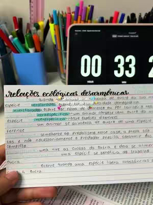The image shows a handwritten study note with highlighted text and a digital timer in the background. The note is in Portuguese, and the timer displays 00:33. There are pens, pencils, and a tablet on the desk, giving a sense of a study environment.