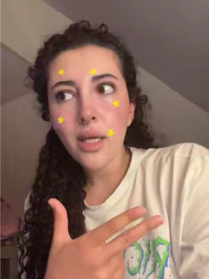 A young woman is centered in the frame, angled slightly towards her left. She has curly dark hair and a septum piercing. Her expression is one of speaking, with her mouth slightly open, and she is glancing towards the left. She is wearing a white t-shirt. Digital yellow stars are on her face. She is gesturing with two fingers. The lighting is soft, illuminating her face. Behind her is a wall.