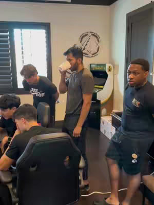 A group of young men are gathered in a room, some standing and some sitting in front of a computer. One man is drinking from a white cup. The room has a window, a pinball machine, and some artwork on the wall. The scene appears to be an educational study session.