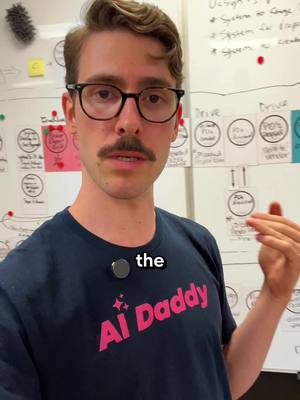 A man with glasses and a mustache stands in front of a whiteboard. The man's outfit includes a navy blue t-shirt with the words "AI Daddy" in pink and white text. A small black circle with the word "the" on it is above the shirt.