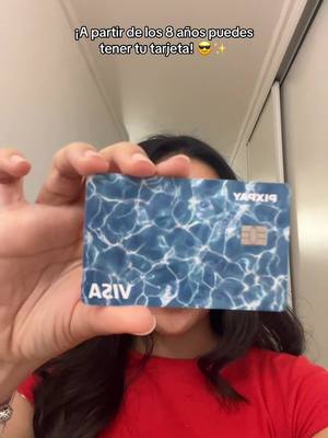 A person is holding a credit card in front of their face. The card is mainly blue with a water ripple design. Text overlaid at the top of the image reads, '¡A partir de los 8 años puedes tener tu tarjeta!'. The person is also wearing a red shirt.