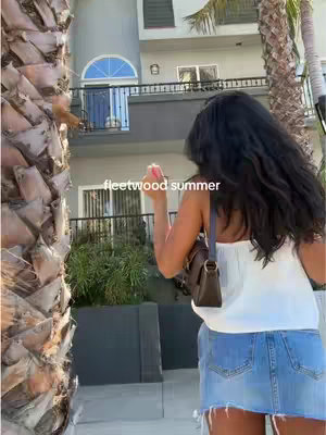 A young woman with long, dark hair is captured from behind against a sunny, outdoor backdrop. She is wearing a white top and a denim skirt. A brown purse hangs over her shoulder. The background features a building with a balcony and arch window partially framed by a palm tree on the left.