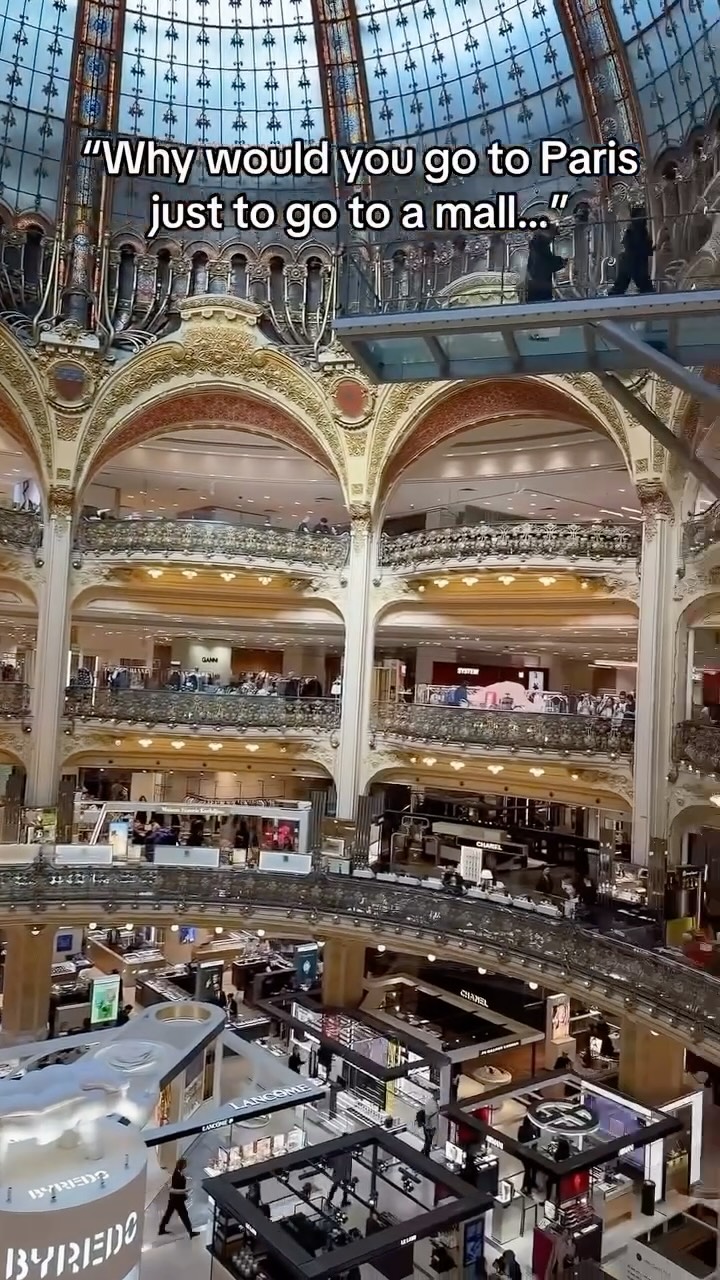 An eye-level view inside the Galeries Lafayette Haussmann department store in Paris. The image is dominated by the store's interior, showcasing multiple levels of shopping floors with ornate decor and a large, stained-glass dome. The architecture is classic and elegant, with gold accents and curved lines.