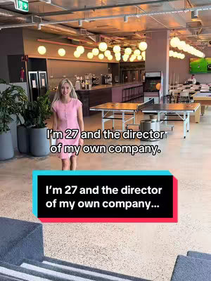 The image showcases a woman standing in a modern office setting. She is facing the camera and smiling with a background that includes office tables and overhead lights. In the foreground, a text overlay sits, with a colorful rectangle.