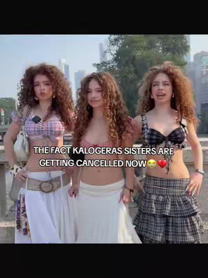 Three young women with long, curly hair stand closely together in front of a blurred cityscape. They are smiling and looking at the camera. Text overlayed in the image reads, "THE FACT KALOGERAS SISTERS ARE GETTING CANCELLED NOW", with sad and heart emojis. The women are dressed in matching crop tops and skirts with different colors and patterns.