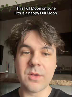 A man with brown hair and a black shirt is the central focus, seen from the chest up. A text overlay is displayed at the top, reading 'This Full Moon on June 11th is a happy Full Moon.' The background features an interior setting with blurred elements like a wall and what appears to be some greenery. The lighting seems natural, and the man's expression is subtly neutral.