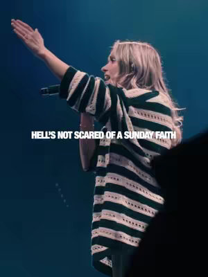 A woman with blonde hair, wearing a black and white patterned top, is singing with one arm raised towards the upper right corner. Her other hand holds a microphone. The background is dark blue with an out-of-focus audience. Overlayed on top is the text 'HELL'S NOT SCARED OF A SUNDAY FAITH' in white.