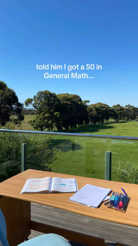 A sunny outdoor scene with a table holding an open book, papers, and markers. The background shows a green field, trees, and a bright blue sky. Text at the top states, 'told him I got a 50 in General Math...'