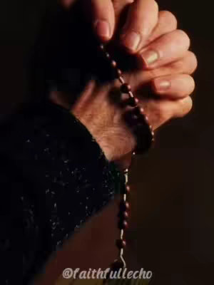 A close-up shot of a pair of hands holding a rosary. The hands are clasped together, with the rosary beads draped between the fingers. The lighting is dim, with a focus on the hands and rosary against a dark background.