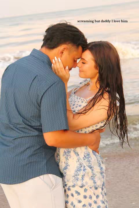 A couple embracing on a beach. The woman is wearing a floral patterned dress, and the man has a button-down shirt. They are touching foreheads, seemingly in an affectionate moment, with an ocean backdrop.
