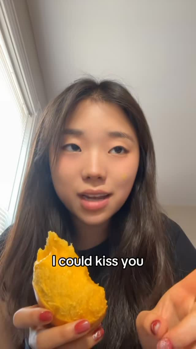 A young woman with long brown hair holds a large piece of fruit, possibly mango, close up. She is indoors, and the background is a light-colored wall. Text overlayed in the image.