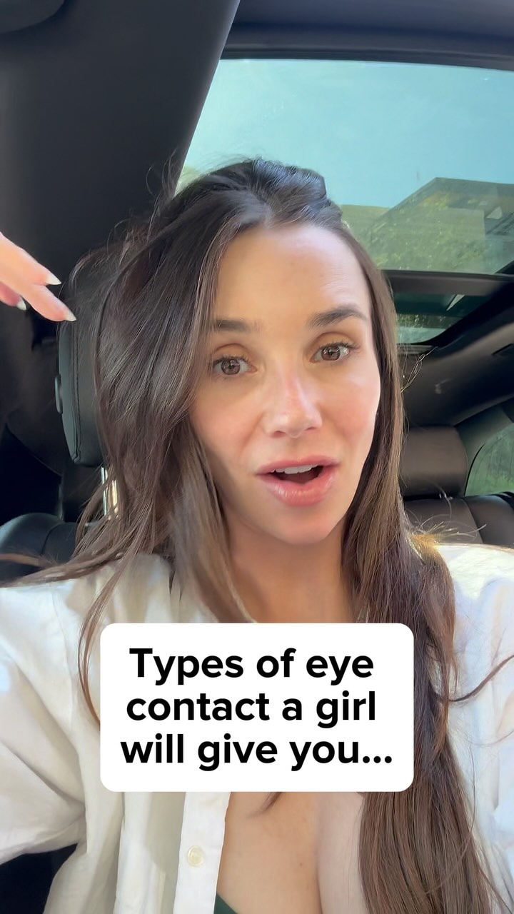 A woman is seated inside a car, facing the camera with a direct gaze and a slight smile. She has long brown hair, and is wearing a white button-down with her neck open. A white rectangular text overlay sits in front of her, which reads 'Types of eye contact a girl will give you...'. The background subtly shows a car interior with a partially visible open sunroof.