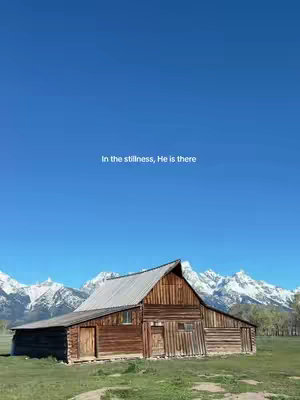 A scenic landscape featuring a rustic wooden barn in the foreground, set against a backdrop of snow-capped mountains and a clear blue sky. The barn is brown with a silver roof and a single door. The mountains dominate the background. The sky is a deep blue. The overall composition evokes tranquility and natural beauty.
