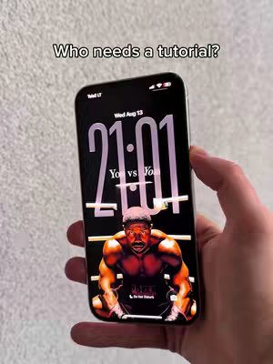 A person holding a black smartphone. The phone's screen displays a Creed movie wallpaper, featuring a muscular black man. Overlayed text asks 'Who needs a tutorial?'. The background is a lightly textured wall.
