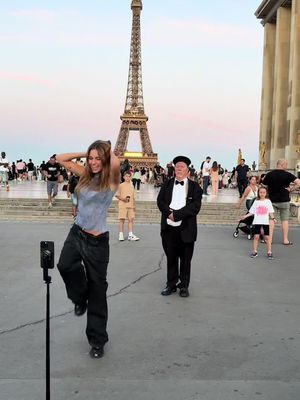 A woman dances in front of the Eiffel Tower, holding her arms up. A man in a tuxedo stands nearby. There are other people in the background.