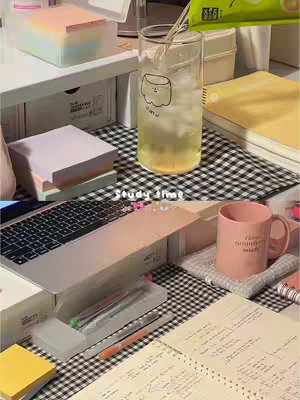 An overhead shot of a desk styled for studying. The desk has a checkered black and white pattern. On the desk are various study-related items: a laptop, sticky notes, books, a glass of iced drink with a straw, a pink mug, pens, and notebooks.