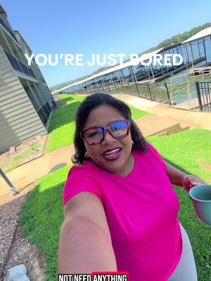 A selfie-style shot of a woman in an outdoor setting. She is smiling at the camera, holding a turquoise mug. She wears glasses with red frames and a pink top. Behind her is a blue sky, green grass, and a body of water with docks and structures.