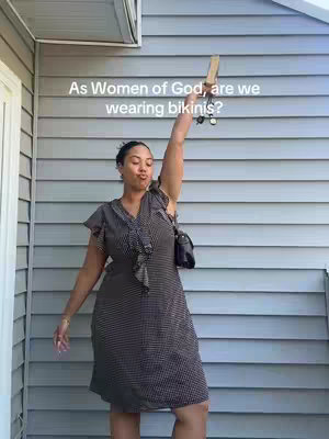 A woman stands in front of a light gray siding background, raising her arm. Text overlay says 'As Women of God, are we wearing bikinis?' The woman is wearing a black and white dress with a black purse. She holds a wooden object in her raised hand. The composition is simple, with the woman centered.