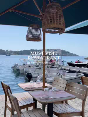 The cover image shows a seaside restaurant in Villefranche-sur-Mer. In the foreground, there are wooden tables and chairs. The background features a view of the sea with boats, a dock, and a coastal landscape. The sky is blue, and the overall color scheme is bright and airy. The text overlay reads 'MUST VISIT RESTAURANT in Villefranche-sur-Mer.'