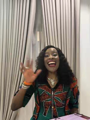 A woman with long, dark curly hair is smiling with her mouth wide open and right hand raised. She has on a colorful patterned top and jewelry. A window with white vertical blinds is visible in the background.