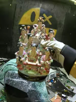 The image shows a smashed car window, with a statue of figurines on a base being held up by a gloved hand. Behind it, a wall has graffiti of a yellow circle and a black X . The scene is poorly lit, giving a sense of destruction in the background.