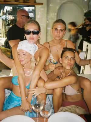 A candid, slightly out-of-focus photo shows four young women seated at a restaurant table, with two embracing and the other two looking towards the camera. The women are in casual outfits, some wearing sunglasses and jewelry. They are in a well-lit restaurant setting, with the background details blurred due to depth of field.