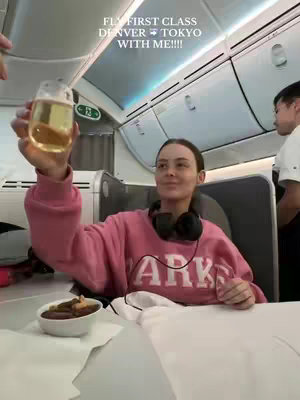 A woman in a pink sweater raises a glass of beer in a first-class airplane cabin. Text overlays indicate the flight is from Denver to Tokyo.