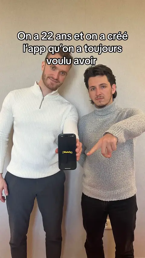 Two men are standing side-by-side, slightly angled towards the camera. The man on the left is wearing a white ribbed, zip-up sweater and the man on the right is wearing a grey turtleneck. The man on the left is holding a phone displaying the 'Mately' app logo at eye level and the man on the right is pointing to the phone with his index finger. Text, positioned at the top, reads, 'On a 22 ans et on a créé l'app qu'on a toujours voulu avoir.' The photo is taken against a plain, cream-colored wall.