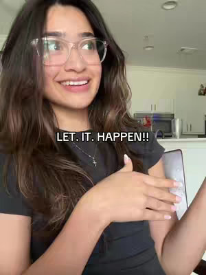 A woman with long, dark hair and glasses smiles while gesturing with her hands. Text overlayed in bold white font reads 'LET. IT. HAPPEN!!'. The background shows a living room.