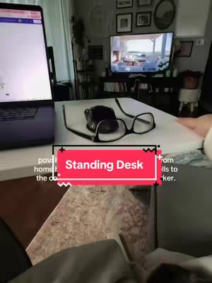 A room is shown with a laptop, glasses, and a computer mouse on a white standing desk. Behind the desk is a tv is displaying a landscape, there are framed photos, white drapes and other decor is also visible.