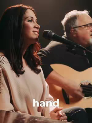 A woman with dark hair is singing into a microphone, while a man plays a guitar in the background. The woman is wearing a light-colored top. The background is dimly lit. The word 'hand' is added as a hook in the lower part of the image.