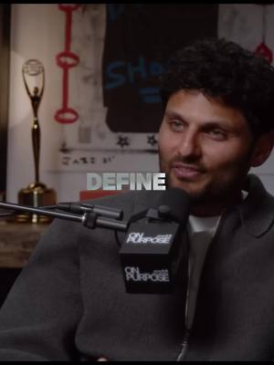 The image is a close-up shot of a man with dark hair and a beard, wearing a dark jacket. He is positioned behind a microphone, likely for a recording or interview. The word "DEFINE" is overlaid in a bold, silver font. There is a background that appears to be a wall with images and decorative elements. The lighting is soft, focusing on the man's face and creating a professional atmosphere.