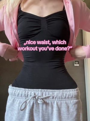 A woman standing in front of a backdrop, wearing a black top over a black camisole, and gray sweatpants. A pink jacket is draped over her shoulders, highlighting her waist, which is emphasized in the image. Text with a pink background overlays the image, asking about the creator's workout routine, which is tied to the fitness-focused nature of the content.
