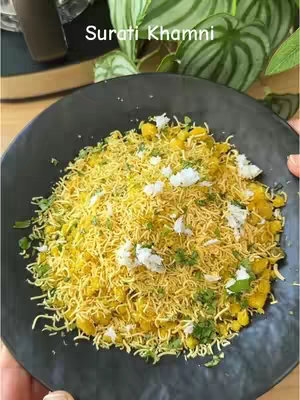 A close-up shot of a plate filled with Surati Khamni, a Gujarati dish. The dish is topped with vibrant yellow sev and garnished with fresh coriander and white grated coconut. The plate is black and set against a slightly blurred background of a tabletop and a green leafy plant.